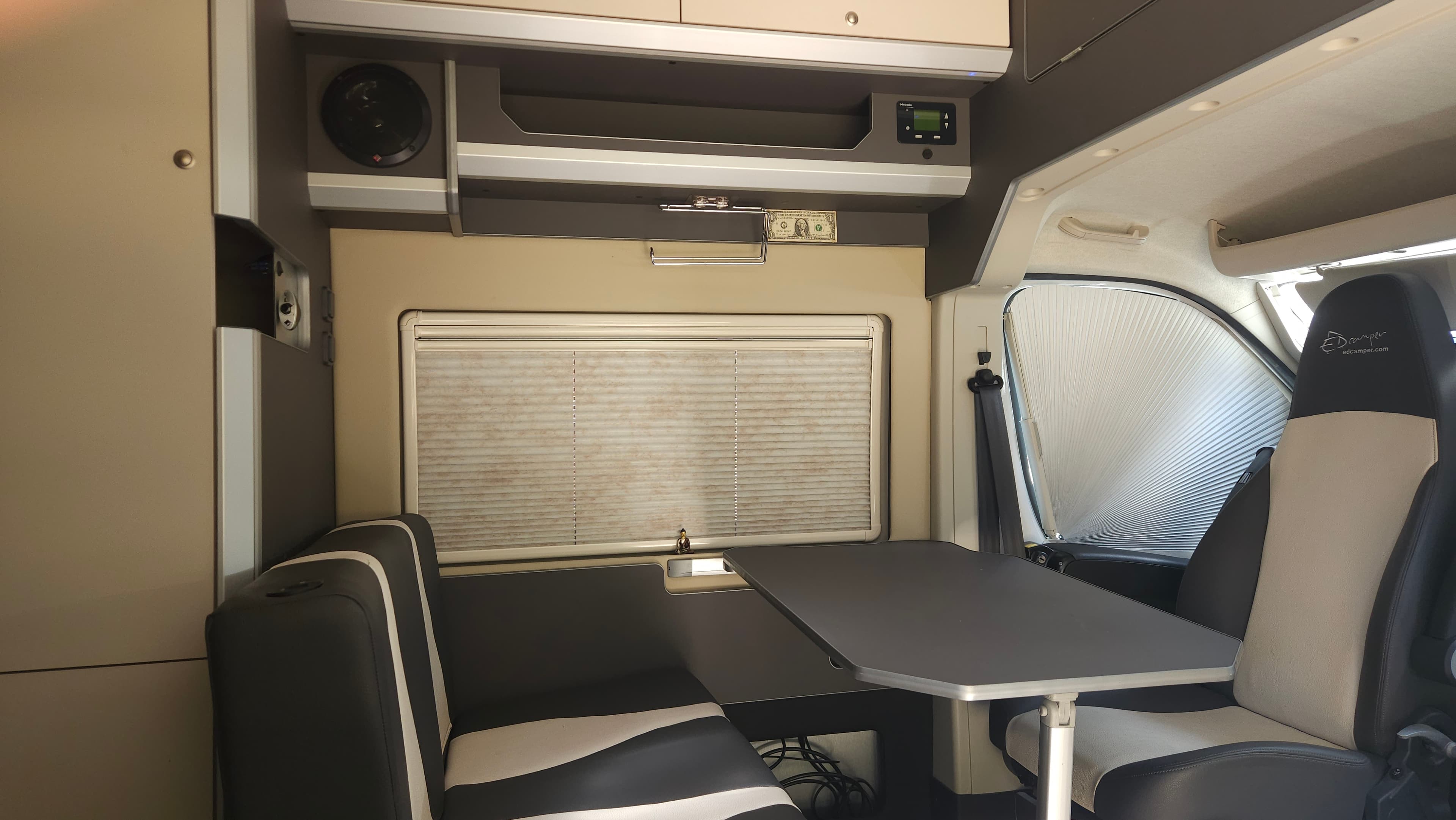 Ventana con persiana bajada; asiento del conductor girado 180° hacia los otros dos asientos (convertibles en cama) y la mesa de comedor. La persiana parece sucia pero esa es su textura: es el único modelo disponible.